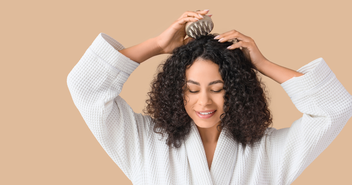 A woman with curly hair is massaging her scalp with a massage brush while wearing a white bathrobe.