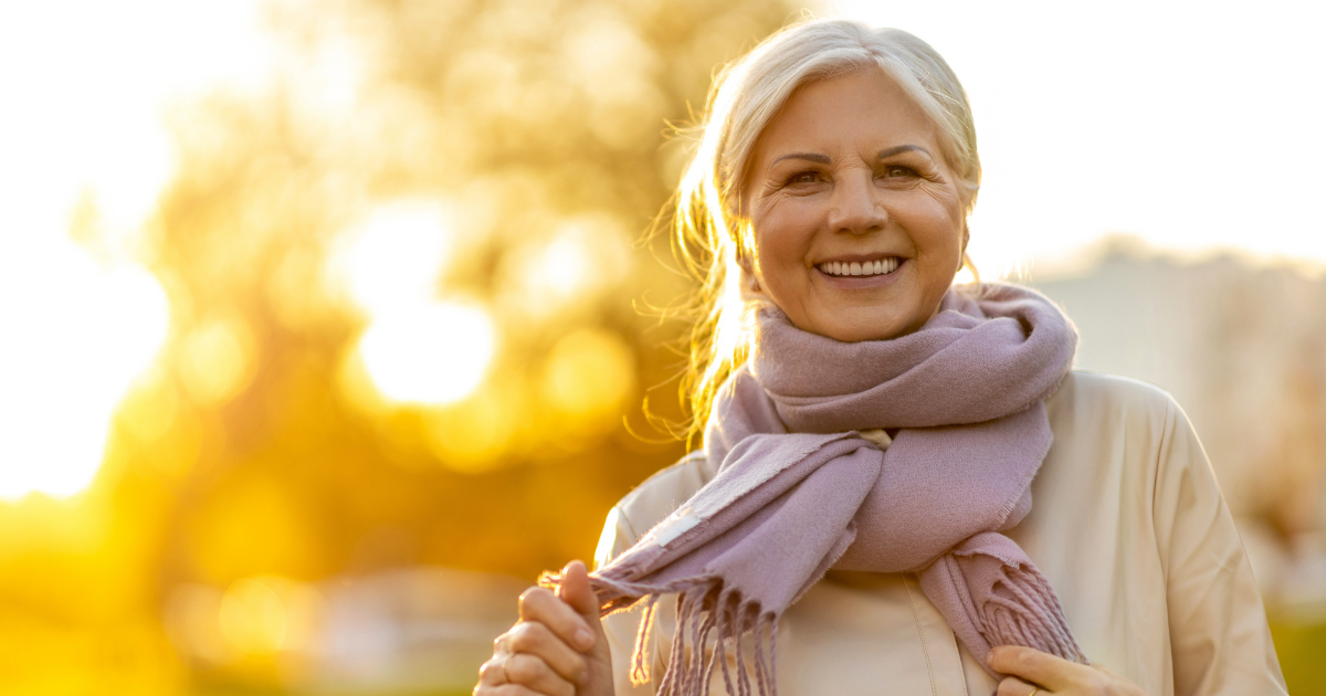 An elderly person wearing a scarf is standing outside in the sunlight, smiling.